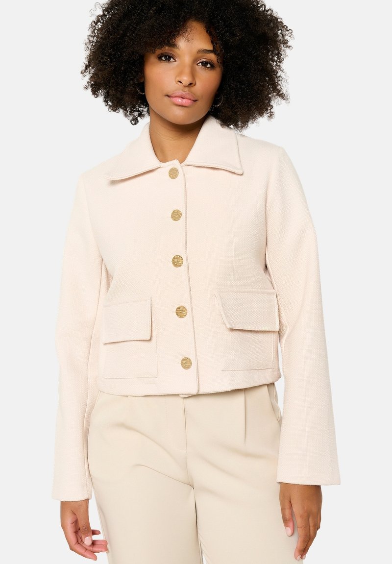 Sagriezta persiku krāsa jaka ar strukturētu audumu, liela apkakle un seši zeltaini pogas, ar divām priekšējām kabatām ar atloka aizdariem.