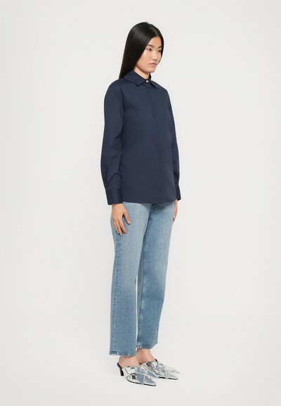 Camicia azzurra con bottoni e colletto, abbinata a jeans azzurri a gamba dritta e scarpe argentate a punta con tacco basso.
