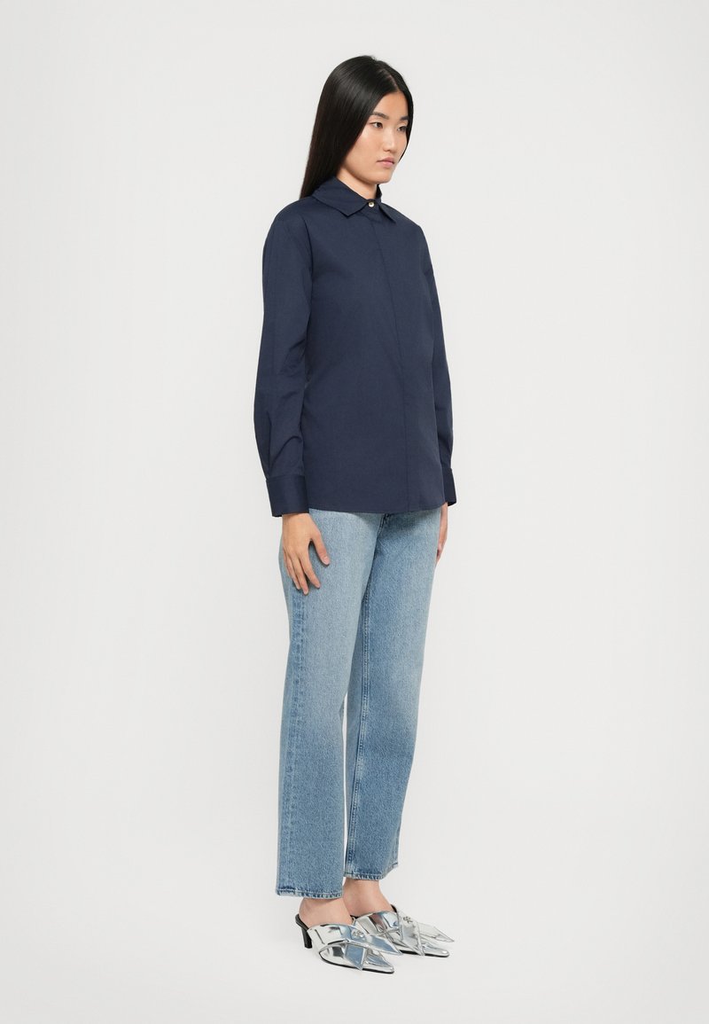 Chemise bleu marine à boutons avec un col, associée à un jean droit bleu clair et des chaussures à talons aigus argentées.