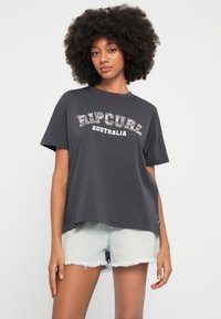 Sötétszürke pamut póló, amelyen világos betűkkel az "RIPCURL AUSTRALIA" felirat olvasható az elején, világos farmer rövidnadrággal párosítva.