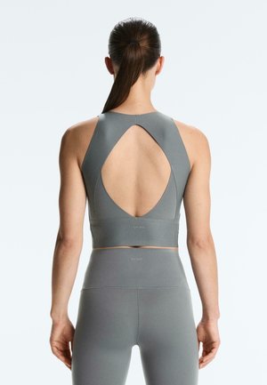 Ženska z rjavimi lasmi, spetimi v čop, oblečena v sive legice z visokim pasom in ujemajočo športno modrček z velikim hrbtnim izrezom v obliki kapljice.