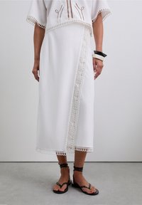 Top court en lin blanc avec des accents en dentelle, associé à une longue jupe blanche ornée d'une bordure en dentelle complexe. Des sandales noires à fines lanières complètent l'ensemble.