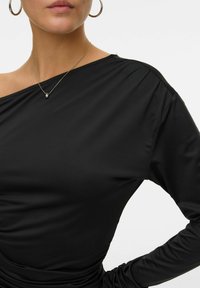 Črna majica z dolgimi rokavi z asimetričnim ovratnikom, mehko teksturo in nagubanim detajlom ob strani, v kombinaciji z srebrnim obeskom.