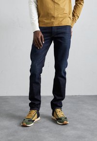 Jeans de mezclilla azul oscuro, ajustados, combinados con zapatillas deportivas verdes y negras con detalles en amarillo. Persona de pie, con las manos relajadas.