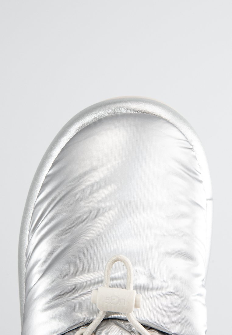 Vista dall'alto di un boot UGG metallico argentato con tessuto imbottito e lacci elastici con toggle su uno sfondo neutro.