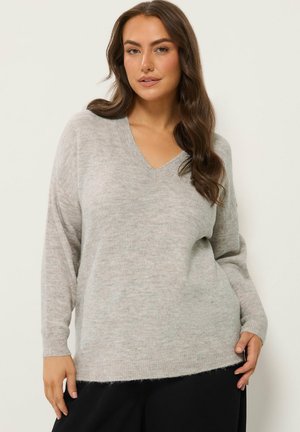 Femme aux longs cheveux bruns, portant un pull gris clair à col en V et un pantalon noir, debout devant un fond clair uni.