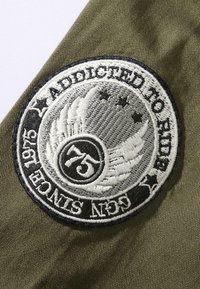 Brodert merke med et sirkulært design med teksten "Addicted to Ride", sølv- og svartfarger, vinger, stjerner og nummeret "75" på olivengrønn bakgrunn.