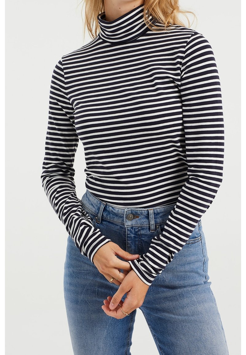 Svart och vitrandig långärmad turtleneck-top med åtsittande design, mjukt tyg, hög krage och kontrasterande randiga manschetter. Blå denimjeans.