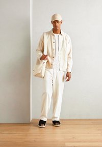 Giacca beige con bottoni sopra una maglietta bianca, abbinata a pantaloni bianchi e sandali neri. Un cappellino beige completa il look, insieme a una borsa in tela.