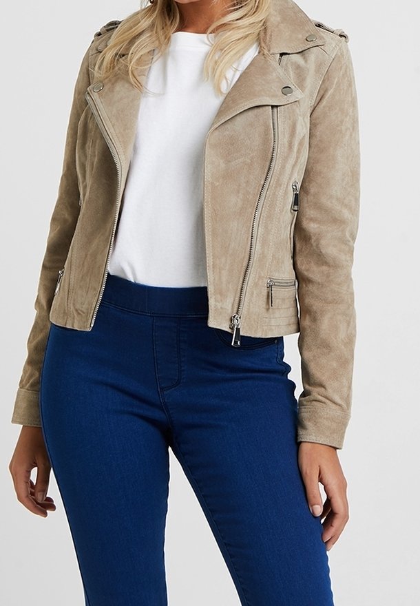 Vrouw draagt een beige suède bikerjack over een wit shirt en donkerblauwe hoge taille spijkerbroek, staand tegen een effen achtergrond.