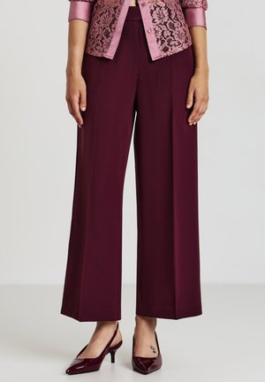 Donna che indossa pantaloni larghi bordeaux, décolleté in vernice coordinati e una blusa di pizzo rosa con bottoni, in piedi su uno sfondo neutro.