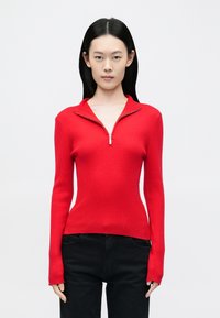 Piros bordázott pulóver negyed cipzáras gallérral, testhez simuló szabással, hosszú ujjakkal és puha textúrával, fekete nadrággal párosítva.