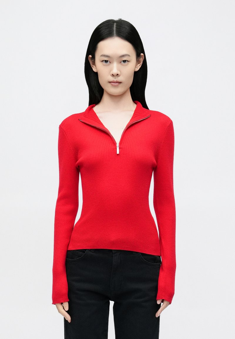 Piros bordázott pulóver negyed cipzáras gallérral, testhez simuló szabással, hosszú ujjakkal és puha textúrával, fekete nadrággal párosítva.