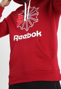 Sudadera roja con capucha que presenta un gran logo blanco de "Reebok" y un diseño geométrico en blanco y rojo. Tiene un bolsillo frontal y capucha con cordones.