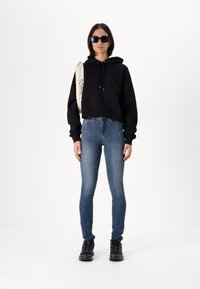 Svart kort hoodie, blå slim-fit jeans och svarta sneakers. Modellen håller en beige axelväska med "TOMMY" tryckt på. Solglasögon närvarande.