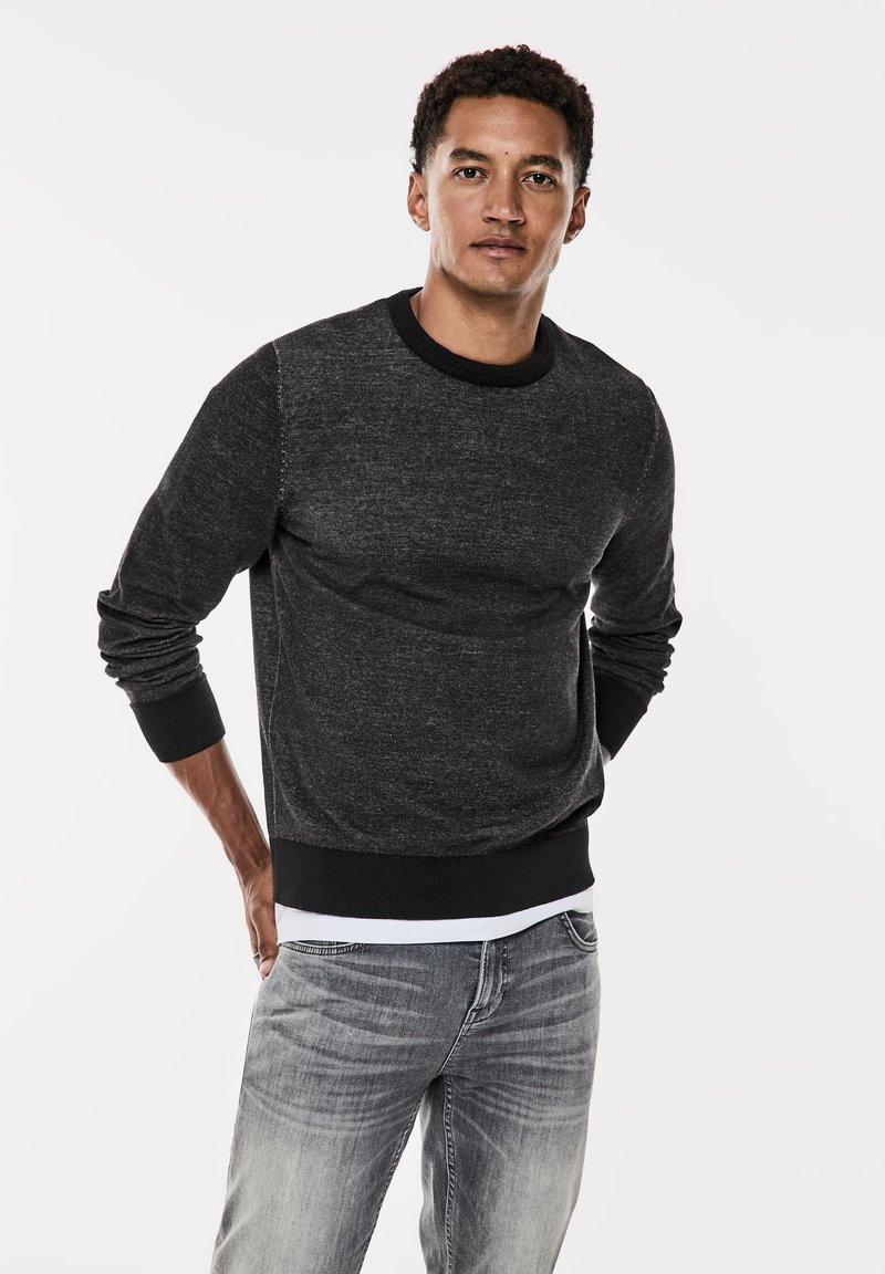 Tmavě šedá mikina s kulatým výstřihem, ribbovaným límcem a manžetami, s měkkou texturou a uvolněným střihem. V kombinaci se světle šedými džíny.