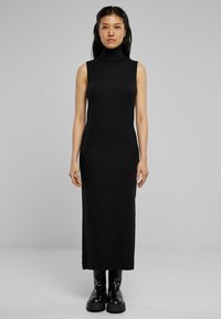 Robe noire sans manches à col montant, silhouette ajustée, mi-longue, en tissu extensible. Portée avec des bottes noires épaisses, se tenant devant un fond uni.