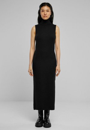 Black, sleeveless turtleneck dress, fitted silhouette, mid-length, made from stretchy fabric. Worn with chunky black boots, standing against a plain background.
