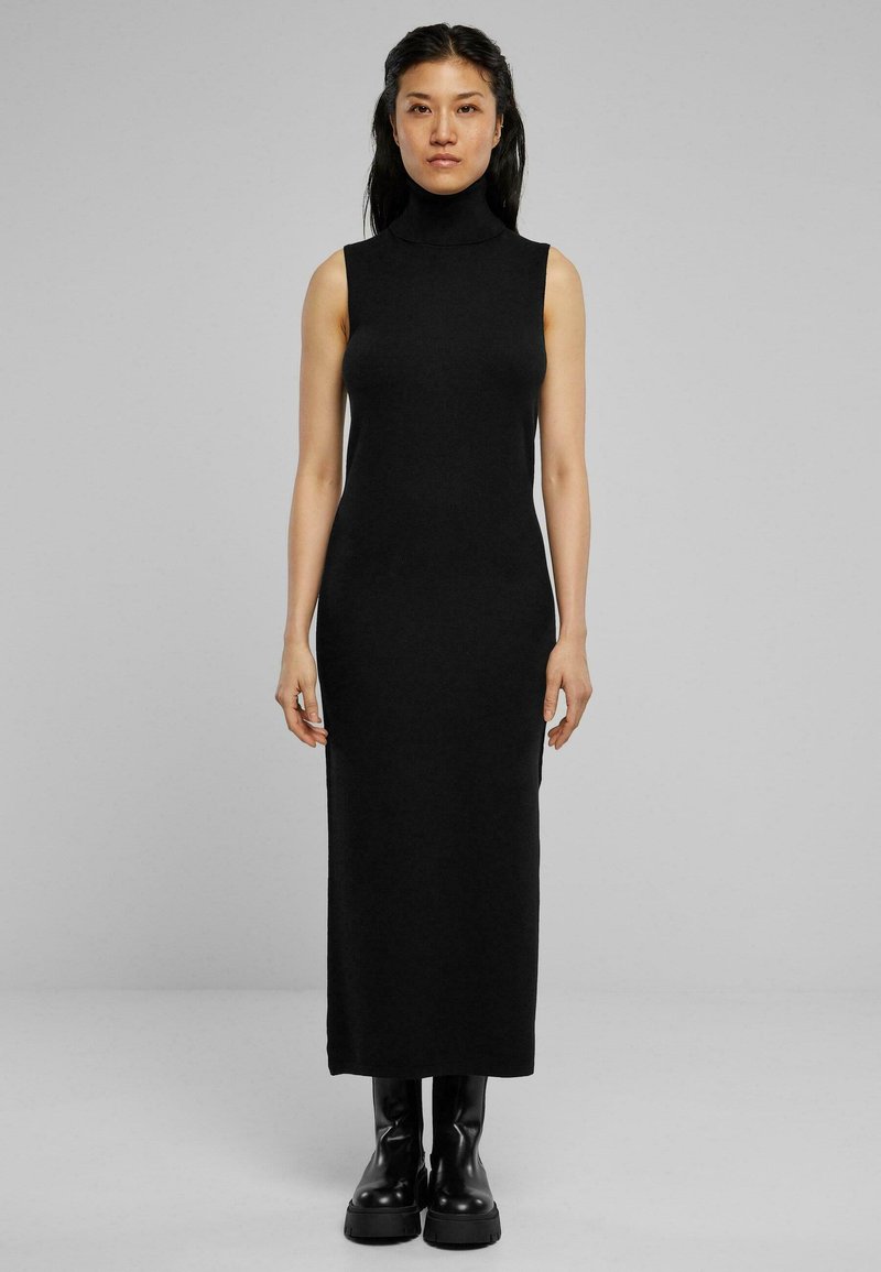 Robe noire sans manches à col montant, silhouette ajustée, mi-longue, en tissu extensible. Portée avec des bottes noires épaisses, se tenant devant un fond uni.