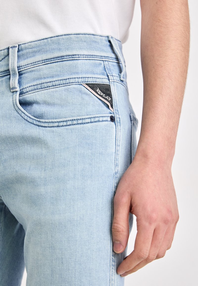 Lichtblauwe denim jeans met een taps toelopende pasvorm, voorzien van een zwart logo patch op de voorzak en subtiele stikseldetails.