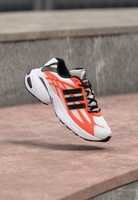 Athletic sneaker in white, orange, and black. Features a textured upper, prominent branding, and a cushioned sole with patterned tread.