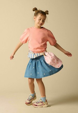 Jong meisje met dubbele knotjes die ronddraait in een roze top met pofmouwen, blauwe denim rok met madeliefjes, sneakers, gestreepte heuptas en roze hoed met hartjesprint.