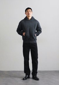Sudadera con capucha de color gris oscuro y un bolsillo frontal, combinada con pantalones negros y zapatos negros. El modelo está de pie frente a un fondo neutro.