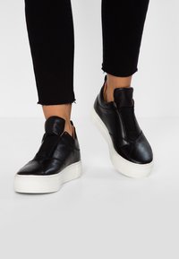 Svarta lädersneakers utan snörning med en vit gummisula, med elastiska sidopaneler och en slät yta.