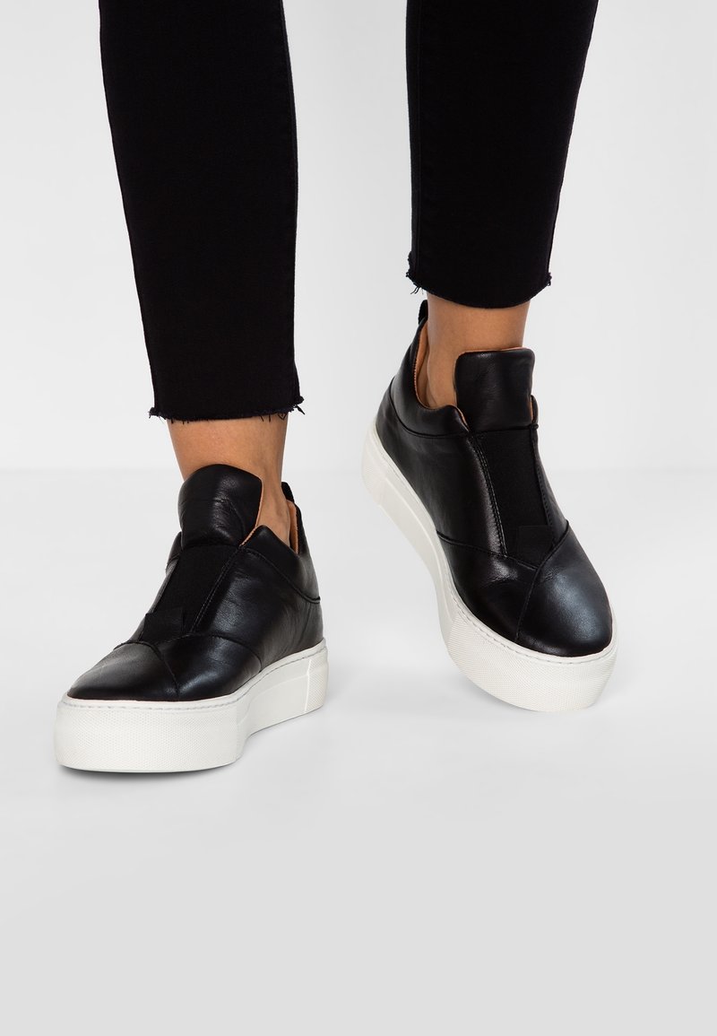 Svarta lädersneakers utan snörning med en vit gummisula, med elastiska sidopaneler och en slät yta.