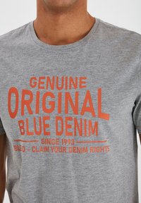 Camiseta de algodón gris con texto en negrita de color naranja que dice "GENUINE ORIGINAL BLUE DENIM". Tiene cuello redondo y mangas cortas.