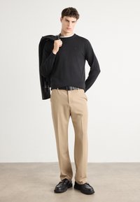 Pull noir à manches longues, pantalon beige et chaussures noires. Un homme tient une veste noire sur son épaule, au design minimal avec des textures discrètes.