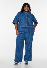 Conjunto de denim en azul oscuro con rayas verticales blancas. Chaqueta corta de manga corta con bolsillo en el pecho, combinada con pantalones de pierna ancha.