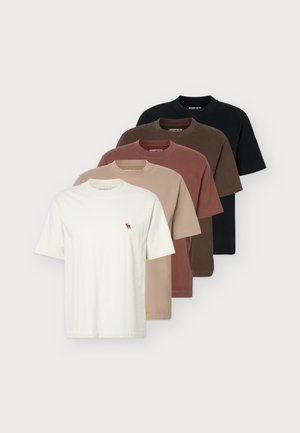 Un homme porte un tee-shirt crème à manches courtes avec un petit logo sur la poitrine, un jean bleu et des chaussures marron sur un fond uni et clair.