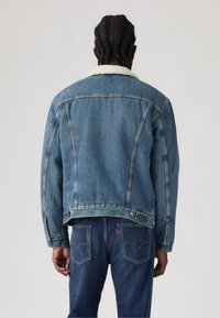 Persona con capelli intrecciati che indossa una giacca di jeans blu con colletto in shearling bianco e jeans blu, di spalle su uno sfondo semplice.