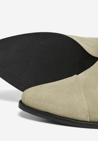 Botines de ante beige de tobillo con punta afilada, que cuentan con una suela de goma negra con textura estriada y un acabado liso y mate.