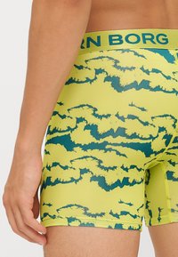 Dzelteni bokseršorti ar tumši zaļām mākoņiem līdzīgām raksta zīmēm, aprīkoti ar zīmolu elastīgu jostasvietu un gludu auduma tekstūru.