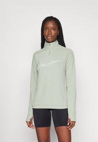 Top de manga larga verde menta con cuello alto y cremallera de cuarto. Presenta un logo blanco en el pecho y un corte ajustado. Se lleva con pantalones cortos negros.