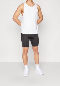 Camiseta deportiva blanca sin mangas y pantalones cortos ajustados negros. El material parece ligero con una textura suave. Zapatillas blancas y calcetines cortos completan el look.