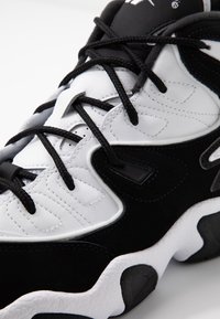 Černobílá kožená teniska s tlustou podrážkou, propracovaným šitím a černými tkaničkami uvázanými přes jazyk.