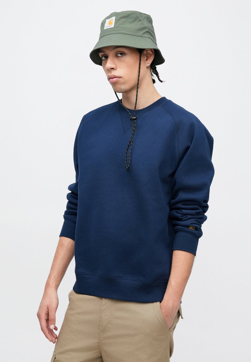 Marineblå sweatshirt med ribkanter ved ærmer og kant, rund halsudskæring og en lille logomærkat. Kombineret med en grøn bøllehat med et logo.