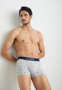 Boxer-briefs en coton gris avec une ceinture marine portant l'inscription "LACOSTE" et de petits motifs en marine. Texture douce, coupe ajustée.