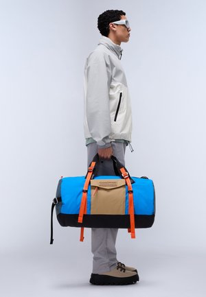 Un sac de voyage bleu et beige avec des anses orange, arborant un patch logo et des accents noirs. Le sac a une forme ronde et est fabriqué en tissu durable.