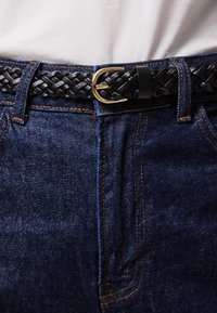Svart flettet lærbelte med spenne i gullfarget metall, plassert over blå denimjeans med oransje søm og glatt tekstur.