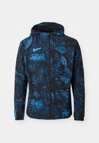 Nike jakke i sort med et blåt abstrakt mønster, lynlås foran, hætte og sidelommer. Fremstillet af letvægtsmateriale for komfort.