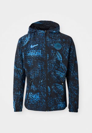 Nike-takki mustana, jossa on sininen abstrakti kuvio, vetoketjulla edessä, huppu ja sivutaskut. Valmistettu kevyestä materiaalista mukavuutta varten.