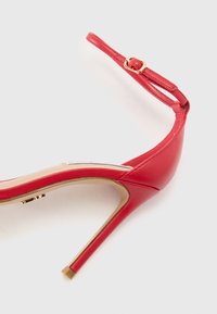 Sandale à talons élevés en cuir rouge avec une fine Bride de cheville, boucle dorée et bout élégant pointu. Surface texturée avec une finition lisse.