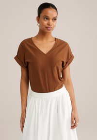 Brązowa bluzka typu v-neck z krótkimi, podwiniętymi rękawami, o teksturowanej tkaninie, w zestawie z białą spódnicą o gładkim wykończeniu i elastycznej talii.