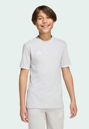 Giovane sorridente che indossa una t-shirt e pantaloni Adidas grigio chiaro, in piedi davanti a uno sfondo chiaro e uniforme.