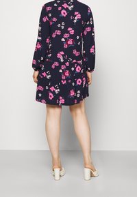 Frau steht mit dem Rücken zur Kamera, trägt ein navyblaues Blumenkleid mit langen Ärmeln und weiße Absatzschuhe mit Slip-On-Design vor einem einfarbigen Hintergrund.