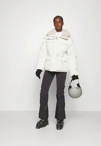 Nainen valkoisessa puffatakkissa, mustissa lasketteluhousuissa, käsineissä ja saappaissa, pitää harmaata laskettelukypärää tyhjää taustaa vasten.